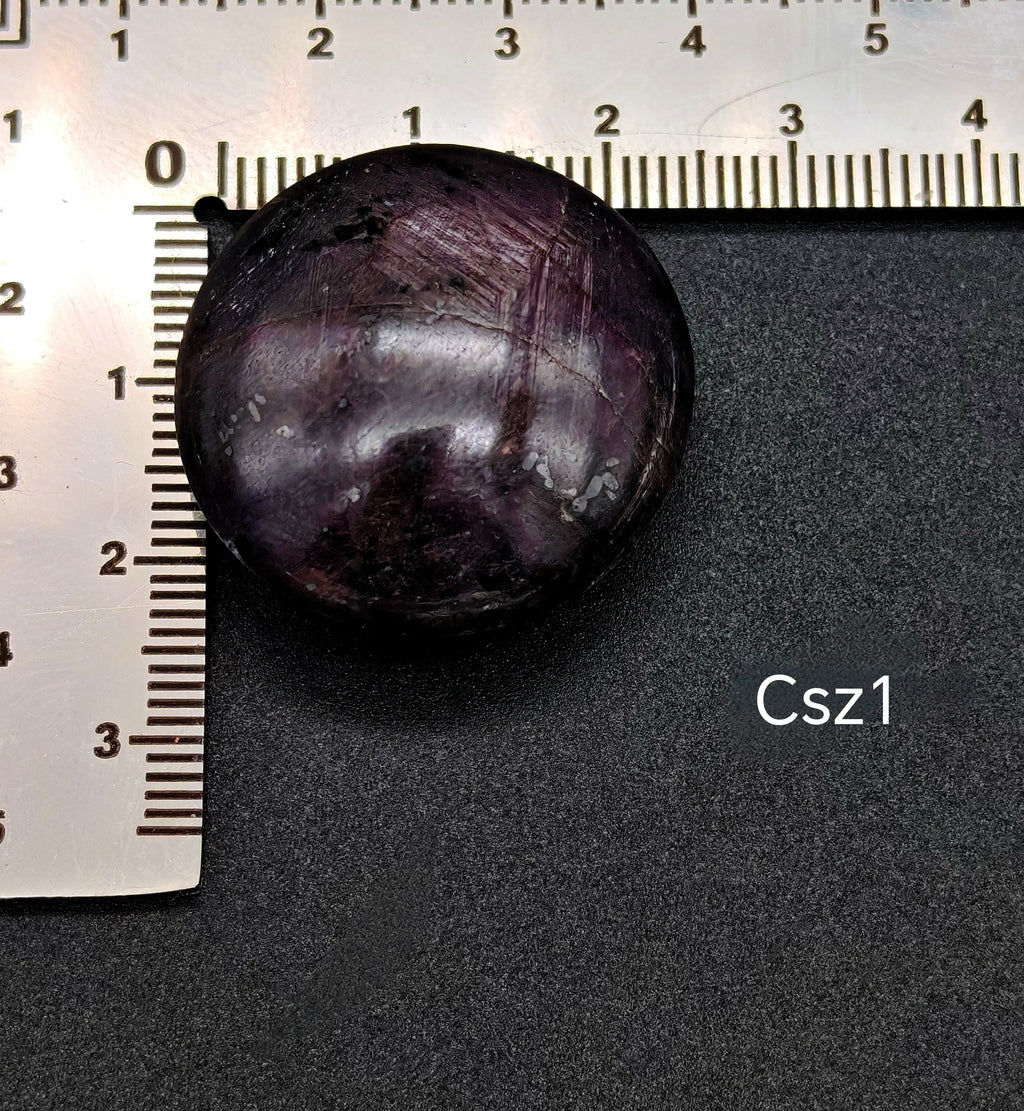 Sötét, lilás árnyalatú csillagzafír kaboson vonalzón mérve, felületén jól látható csillagfénnyel (Csz1)