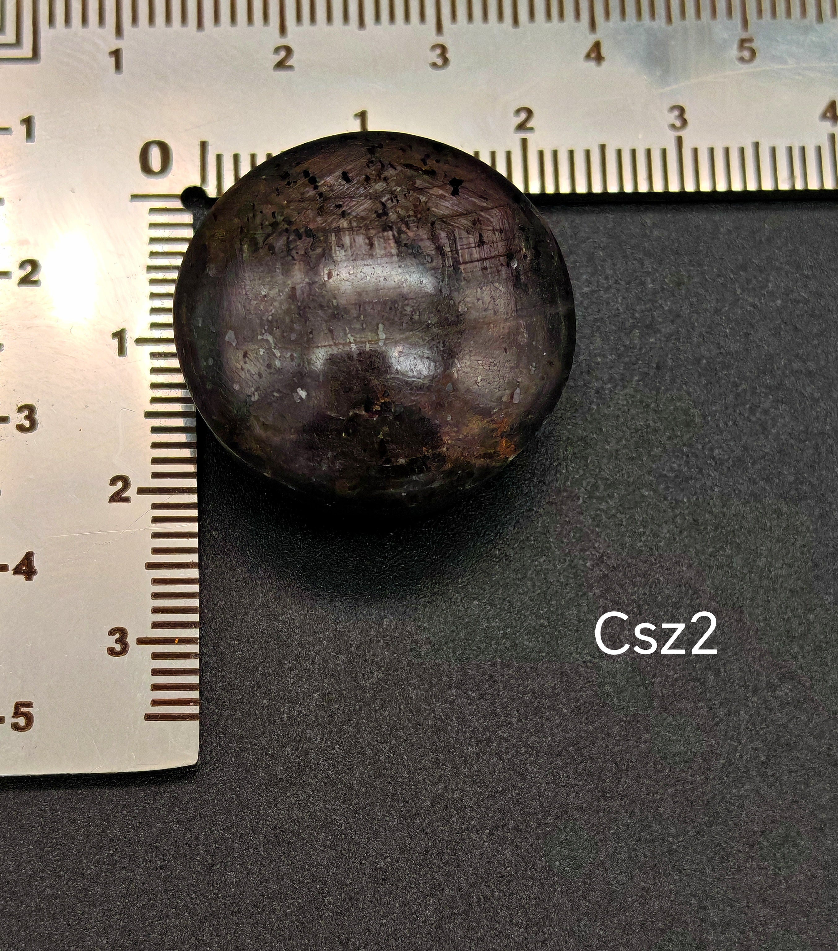 Sötét, lilás árnyalatú csillagzafír kaboson vonalzón mérve, felületén jól látható csillagfénnyel (Csz2)