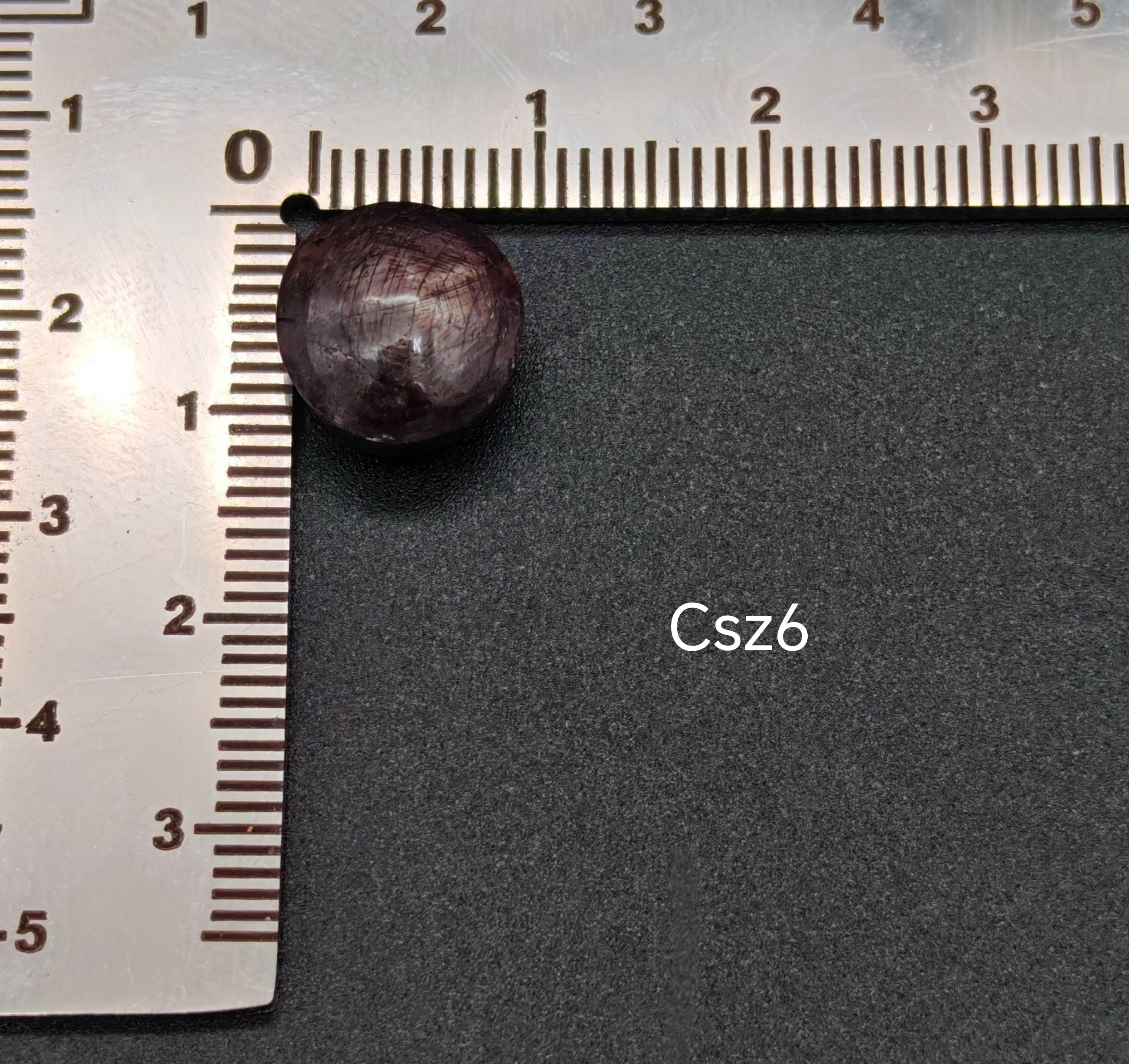 Sötét, lilás árnyalatú csillagzafír kaboson vonalzón mérve, felületén jól látható csillagfénnyel (Csz6)