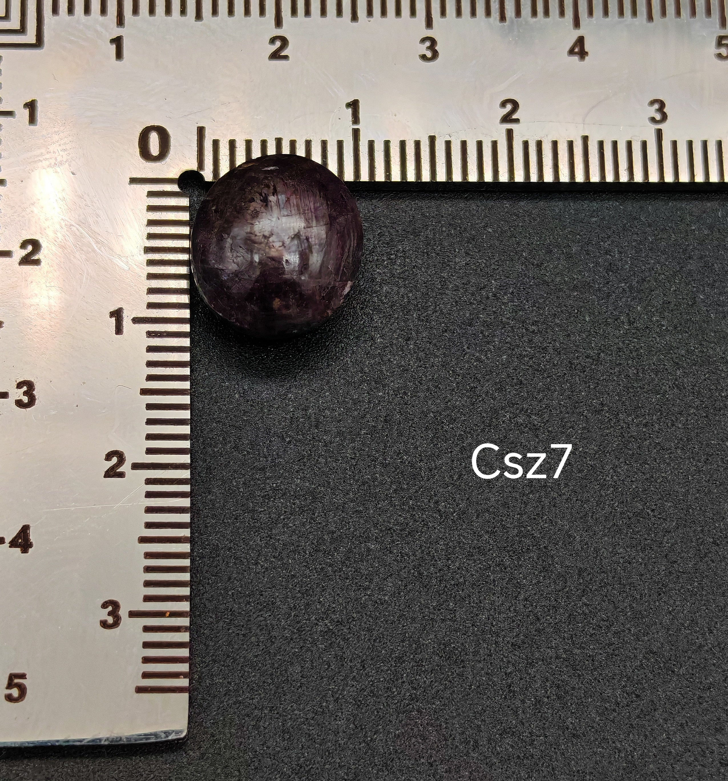 Sötét, lilás árnyalatú csillagzafír kaboson vonalzón mérve, felületén jól látható csillagfénnyel (Csz7)