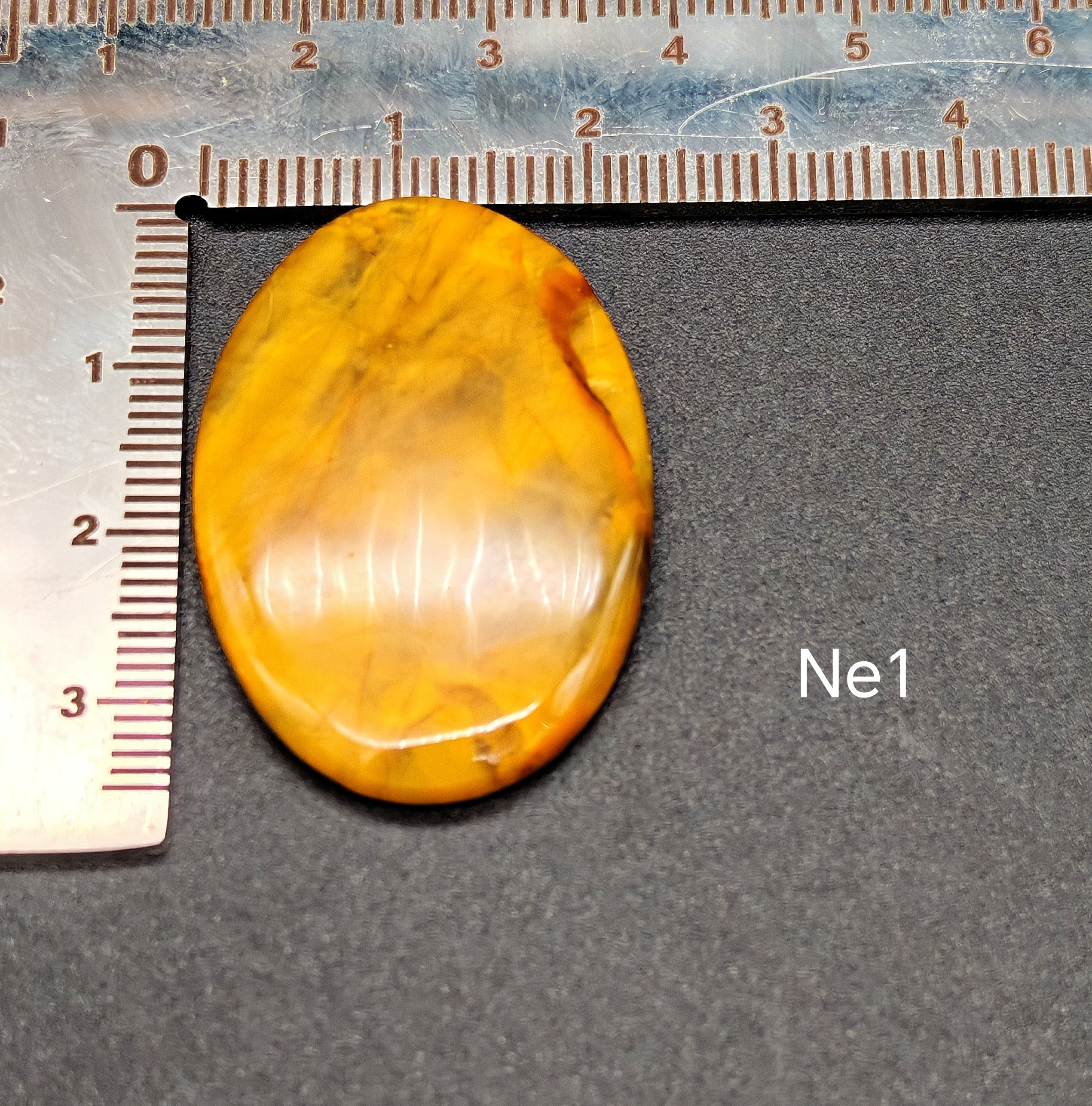 Természetes nellite kaboson meleg borostyán-narancs színben Ne1 jelöléssel, sötét háttéren vonalzóval, mérete kb. 2-3 cm