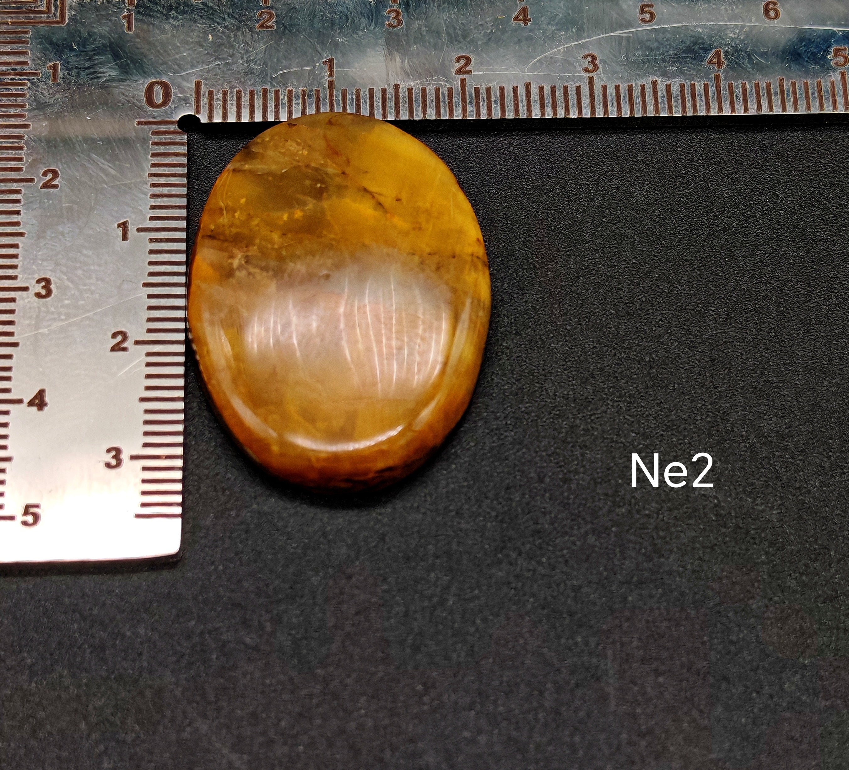 Természetes nellite kaboson meleg borostyán-narancs színben Ne2 jelöléssel, sötét háttéren vonalzóval, mérete kb. 3-4 cm"