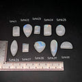 Természetes szivárványos holdkő (rainbow moonstone) kaboson kövek sorban számozva (SZHk21–SZHk30), sötét háttéren vonalzóval, különböző alakokban — a kép sorrendje megegyezik a videóban látható kövek sorrendjével