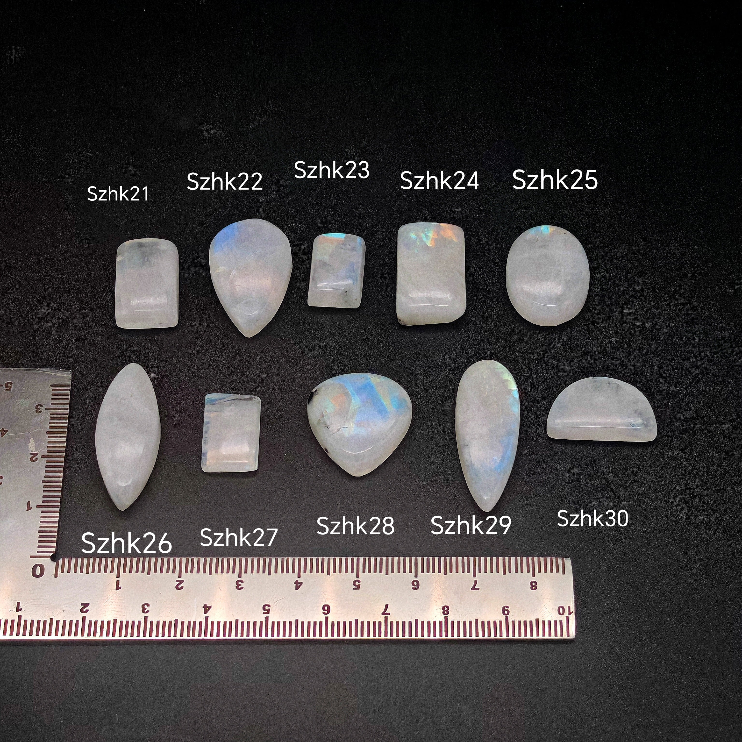 Természetes szivárványos holdkő (rainbow moonstone) kaboson kövek sorban számozva (SZHk21–SZHk30), sötét háttéren vonalzóval, különböző alakokban — a kép sorrendje megegyezik a videóban látható kövek sorrendjével