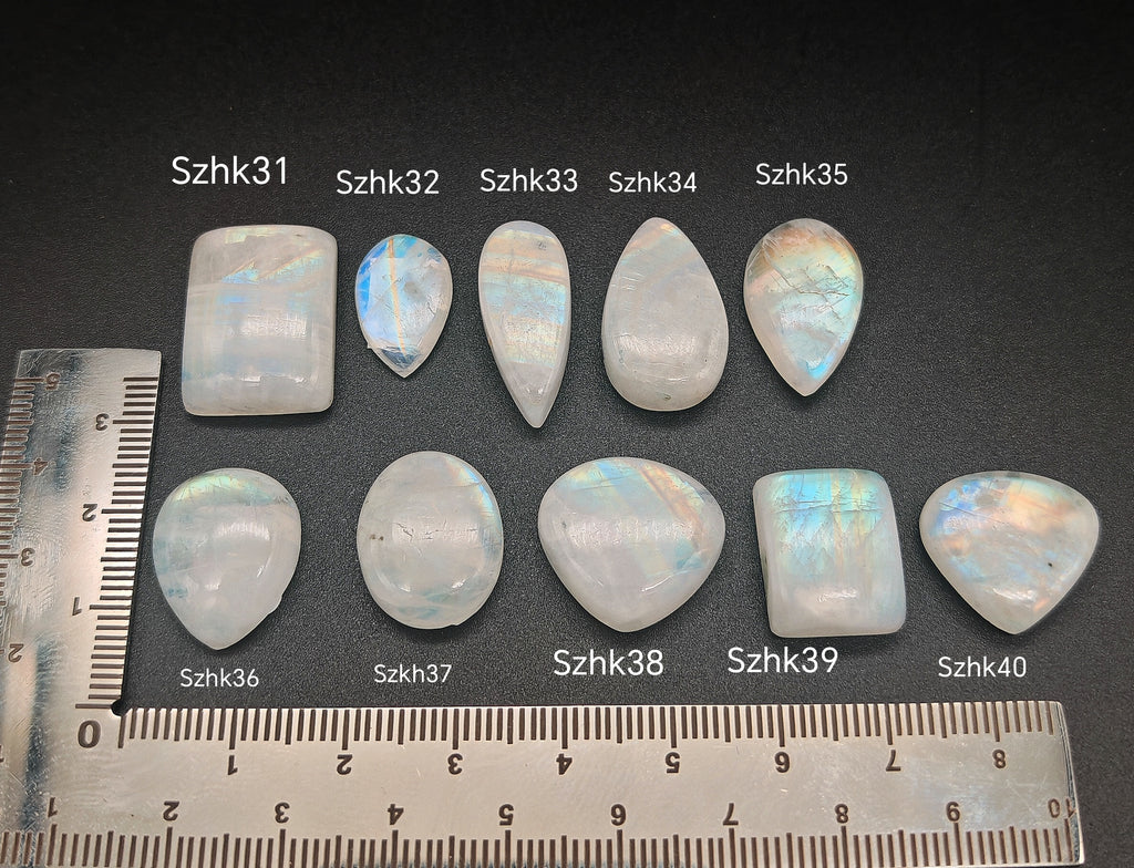 Természetes szivárványos holdkő (rainbow moonstone) kaboson kövek sorban számozva (Szhk31–Szhk40), fehér alapon vonalzóval, különböző alakokban — a kép sorrendje megegyezik a videóban látható kövek sorrendjével