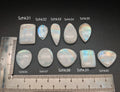 Természetes szivárványos holdkő (rainbow moonstone) kaboson kövek sorban számozva (Szhk31–Szhk40), fehér alapon vonalzóval, különböző alakokban — a kép sorrendje megegyezik a videóban látható kövek sorrendjével