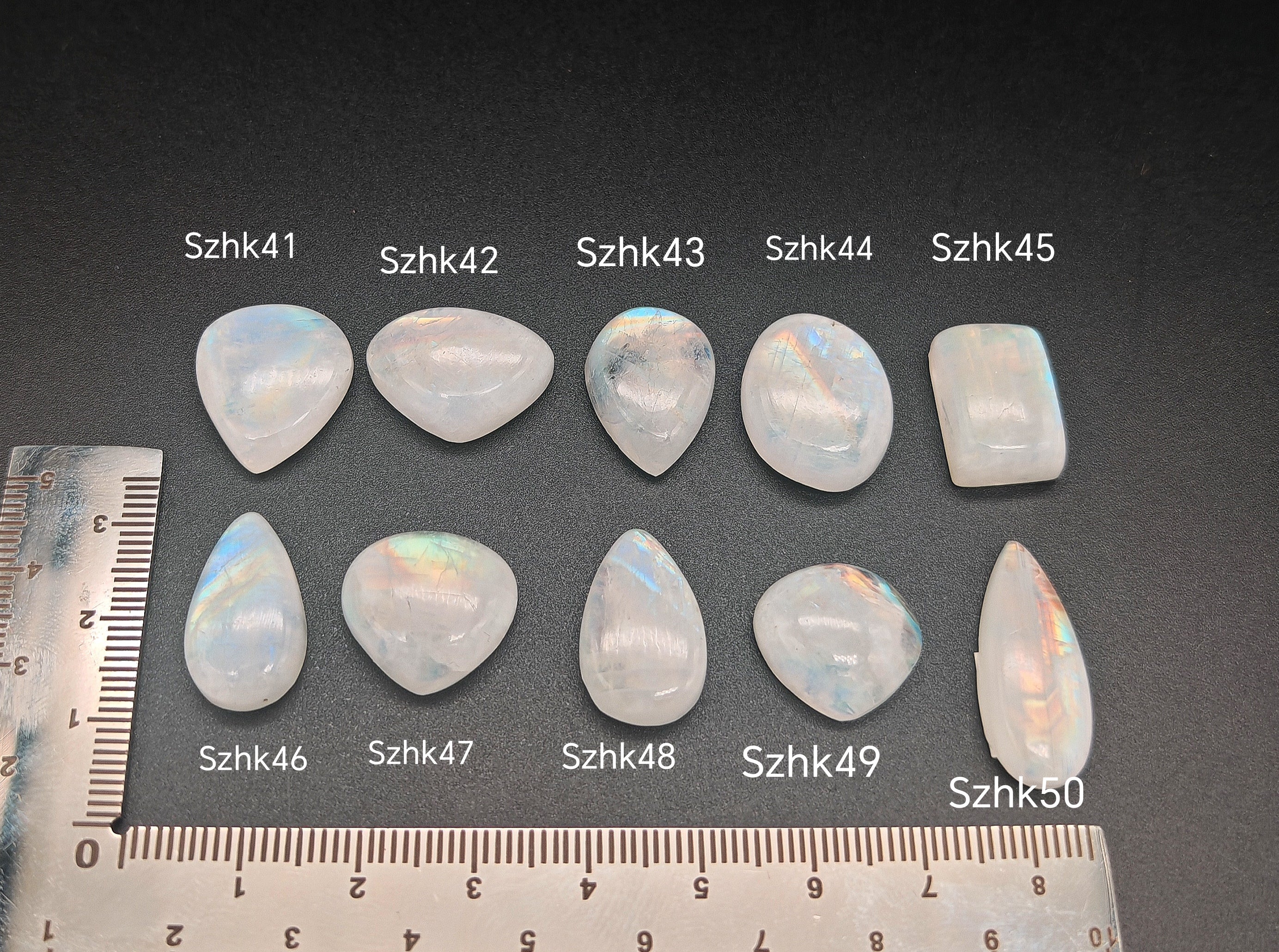 Természetes szivárványos holdkő (rainbow moonstone) kaboson kövek sorban számozva (Szhk41–Szhk50), fehér alapon vonalzóval, különböző alakokban — a kép sorrendje megegyezik a videóban látható kövek sorrendjével