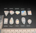 Természetes szivárványos holdkő (rainbow moonstone) kaboson kövek sorban számozva (Szhk51–Szhk60), fehér alapon vonalzóval, különböző alakokban — a kép sorrendje megegyezik a videóban látható kövek sorrendjével