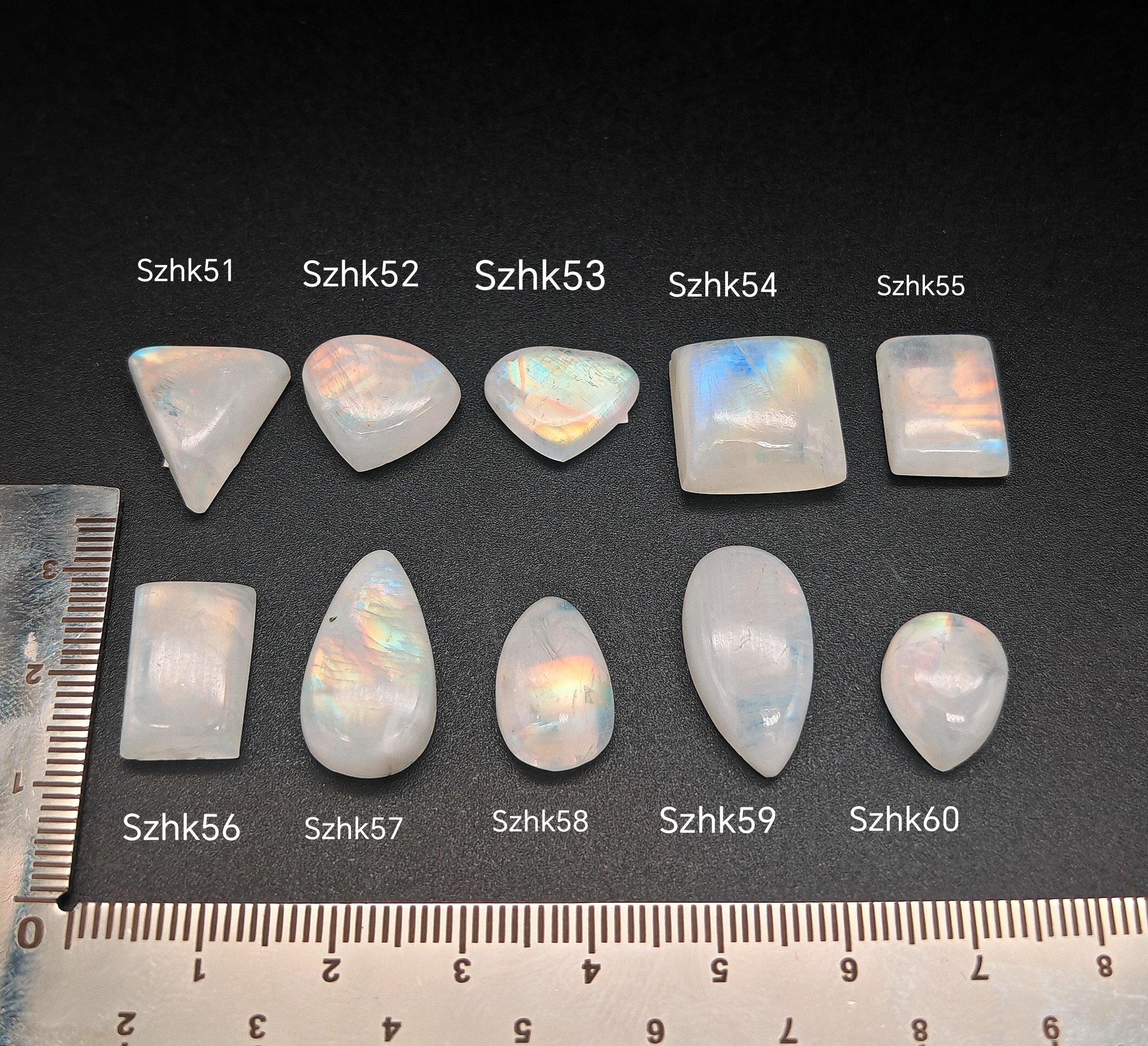 Természetes szivárványos holdkő (rainbow moonstone) kaboson kövek sorban számozva (Szhk51–Szhk60), fehér alapon vonalzóval, különböző alakokban — a kép sorrendje megegyezik a videóban látható kövek sorrendjével