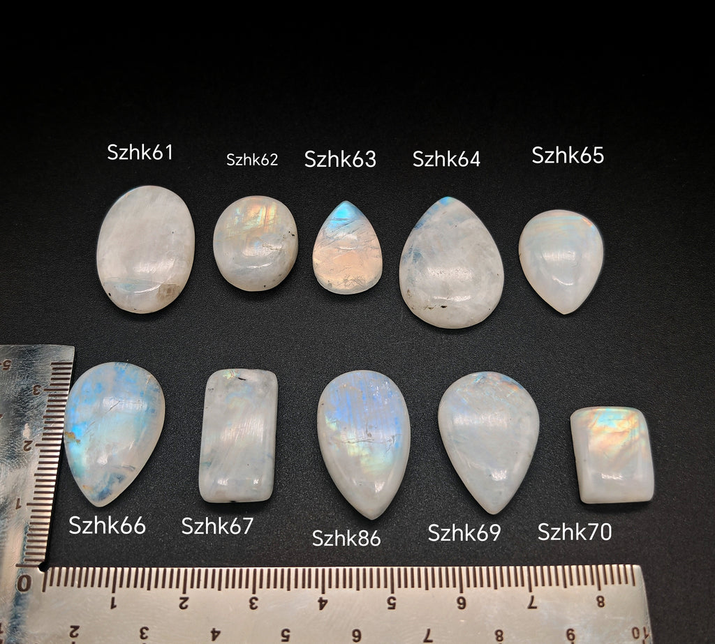 Természetes szivárványos holdkő (rainbow moonstone) kaboson kövek sorban számozva (Szhk61–Szhk70), fehér alapon vonalzóval, különböző alakokban — a kép sorrendje megegyezik a videóban látható kövek sorrendjével