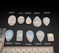 Természetes szivárványos holdkő (rainbow moonstone) kaboson kövek sorban számozva (Szhk61–Szhk70), fehér alapon vonalzóval, különböző alakokban — a kép sorrendje megegyezik a videóban látható kövek sorrendjével
