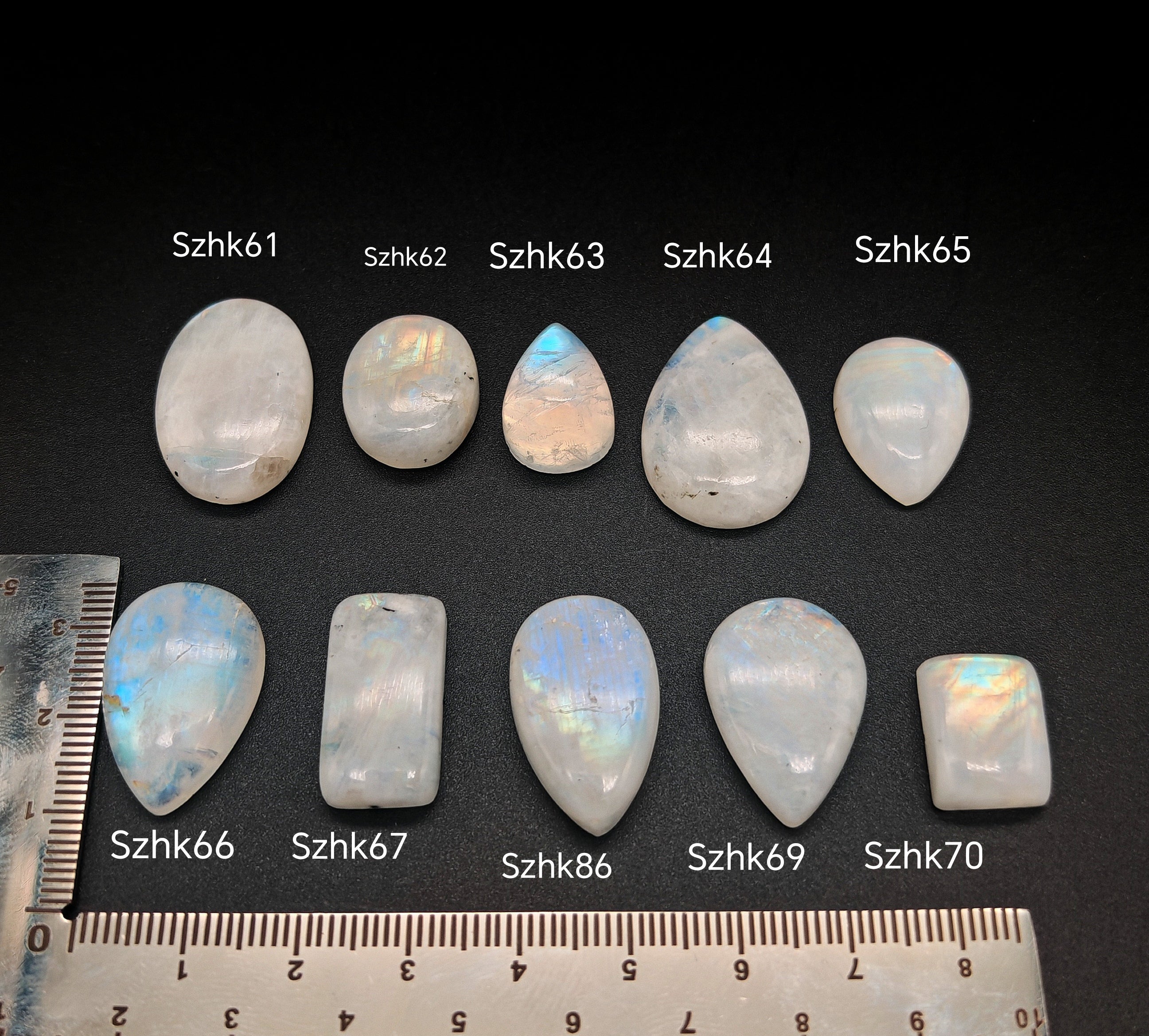 Természetes szivárványos holdkő (rainbow moonstone) kaboson kövek sorban számozva (Szhk61–Szhk70), fehér alapon vonalzóval, különböző alakokban — a kép sorrendje megegyezik a videóban látható kövek sorrendjével