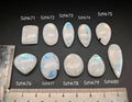 Természetes szivárványos holdkő (rainbow moonstone) kaboson kövek sorban számozva (Szhk71–Szhk80), fehér alapon vonalzóval, különböző alakokban — a kép sorrendje megegyezik a videóban látható kövek sorrendjével
