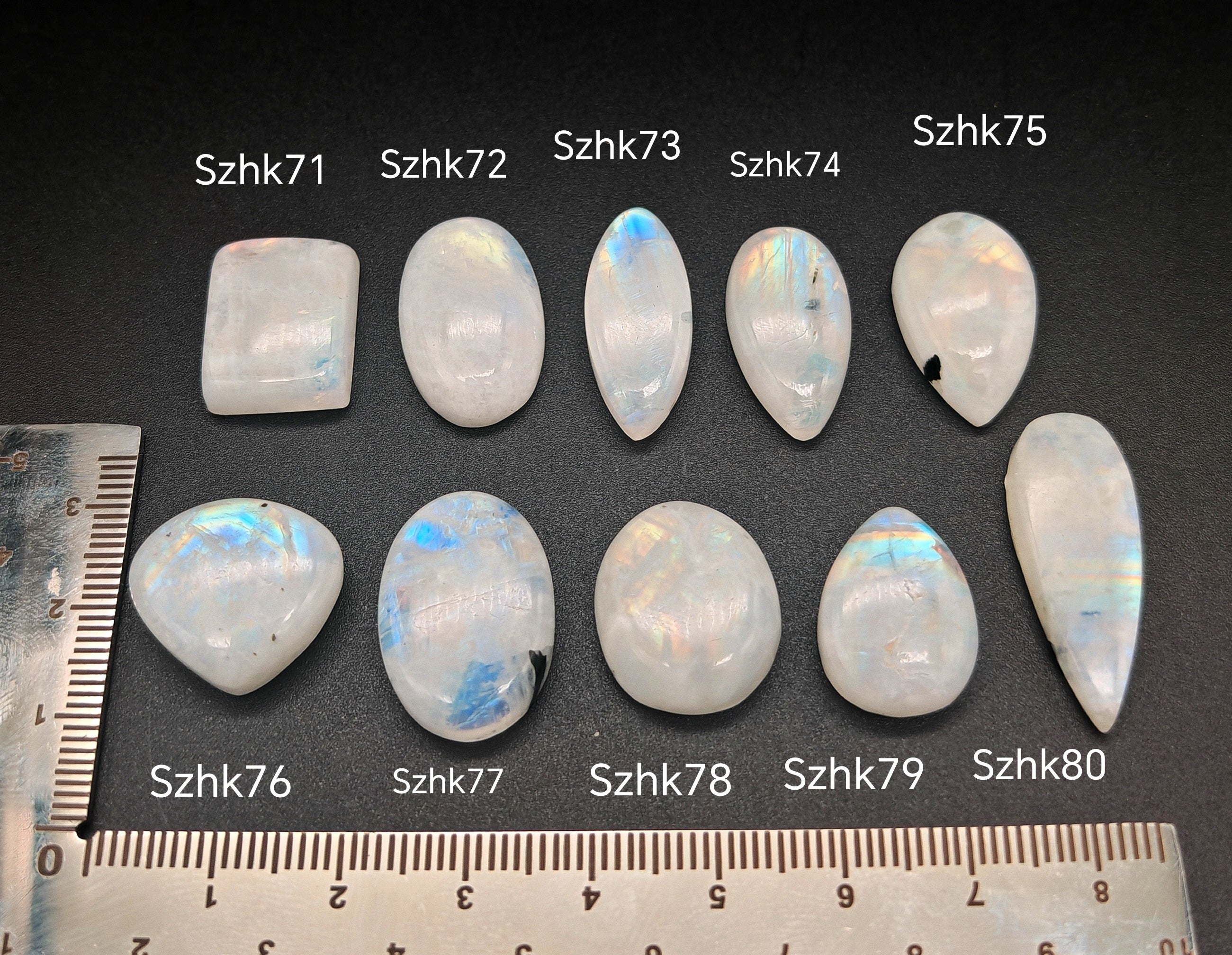 Természetes szivárványos holdkő (rainbow moonstone) kaboson kövek sorban számozva (Szhk71–Szhk80), fehér alapon vonalzóval, különböző alakokban — a kép sorrendje megegyezik a videóban látható kövek sorrendjével