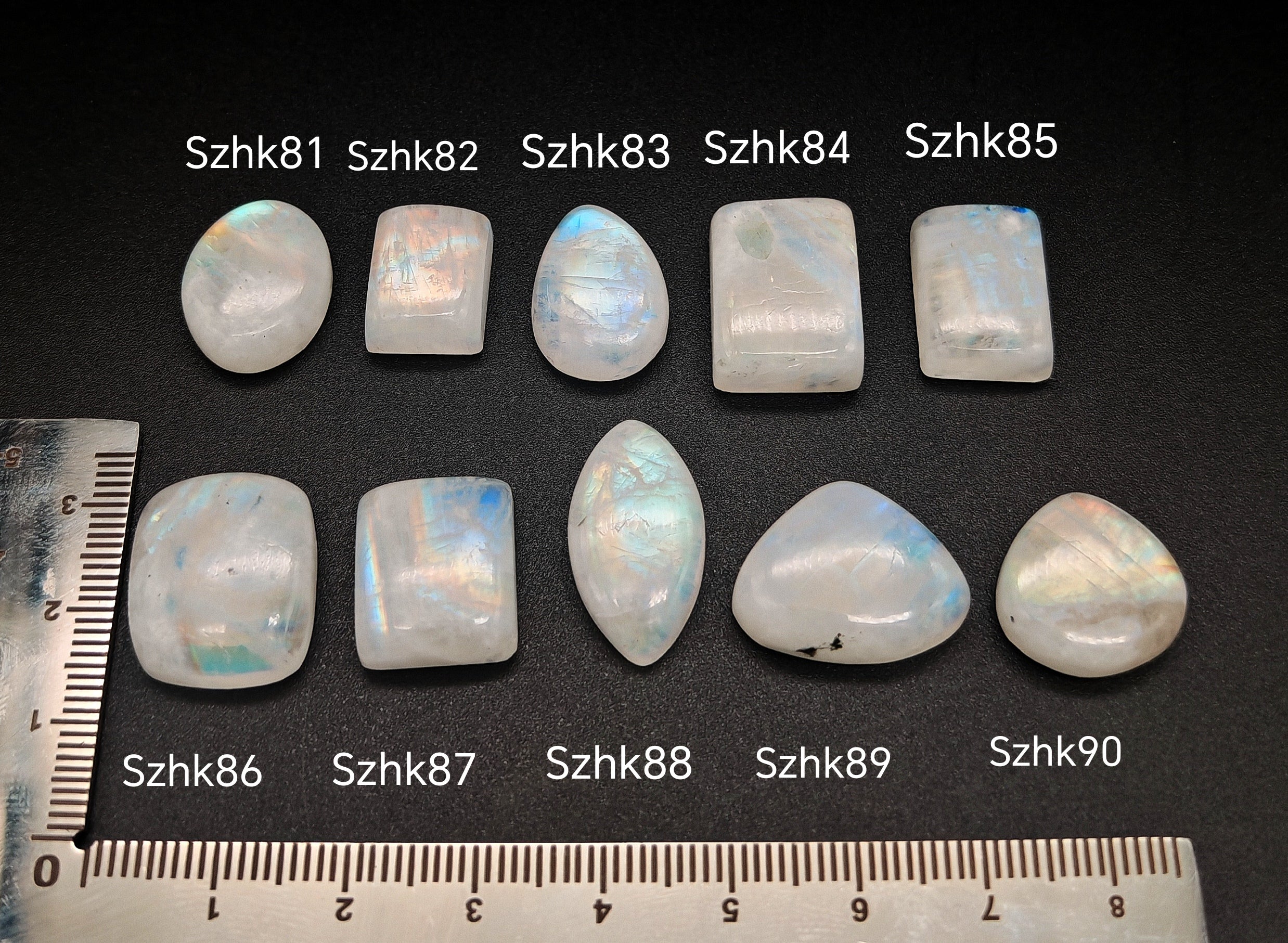 Természetes szivárványos holdkő (rainbow moonstone) kaboson kövek sorban számozva (Szhk81–Szhk90), fehér alapon vonalzóval, különböző alakokban — a kép sorrendje megegyezik a videóban látható kövek sorrendjével