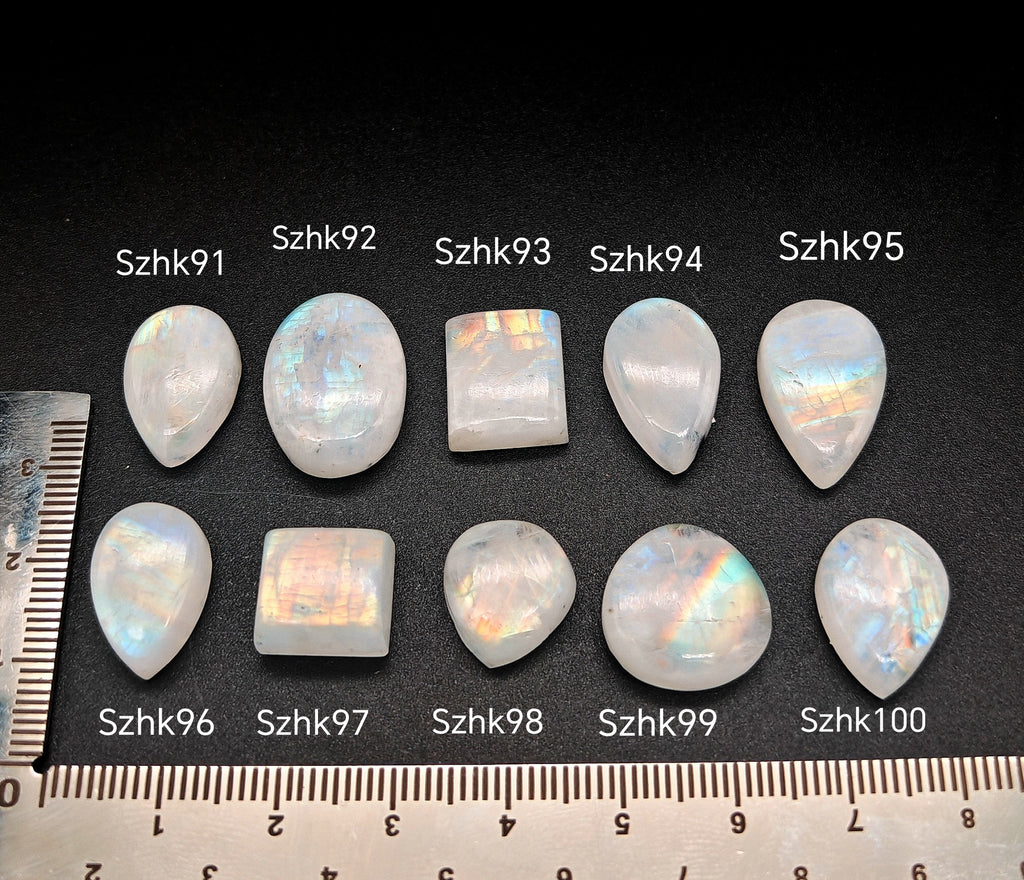 „Természetes szivárványos holdkő (rainbow moonstone) kaboson kövek sorban számozva (Szhk91–Szhk100), fehér alapon vonalzóval, különböző alakokban — a kép sorrendje megegyezik a videóban látható kövek sorrendjével