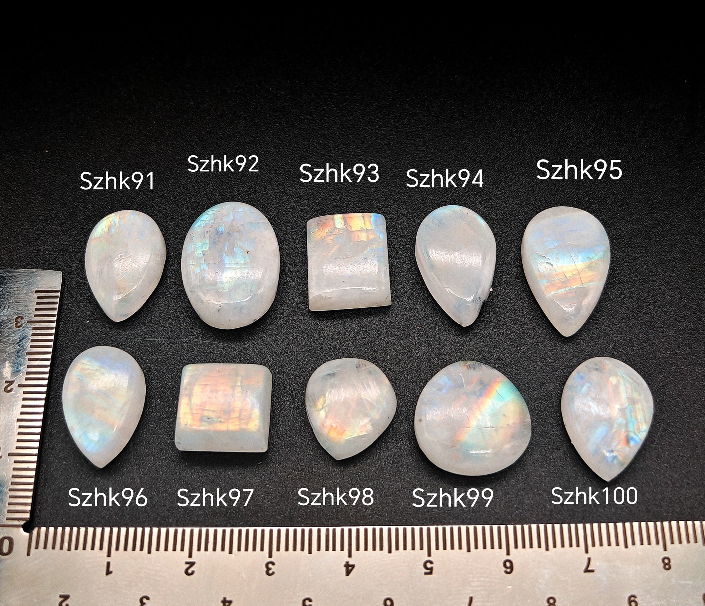 „Természetes szivárványos holdkő (rainbow moonstone) kaboson kövek sorban számozva (Szhk91–Szhk100), fehér alapon vonalzóval, különböző alakokban — a kép sorrendje megegyezik a videóban látható kövek sorrendjével