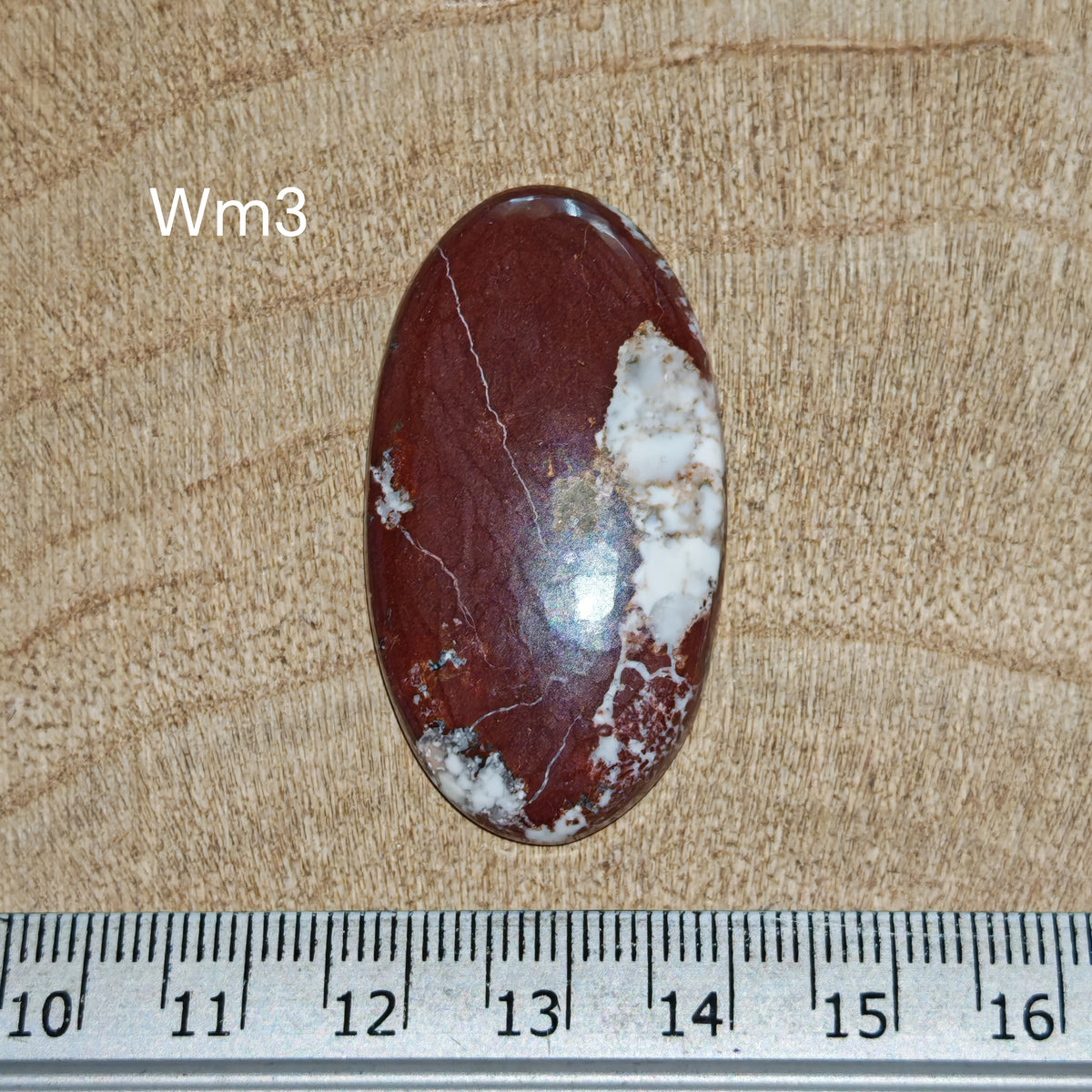 Wildhorse magnezit kaboson, Wm3, ovális formájú, erőteljes vörösesbarna természetes résszel, mellette világos fehér–szürkés, mátrixmintás területtel, milliméteres vonalzó mellett.