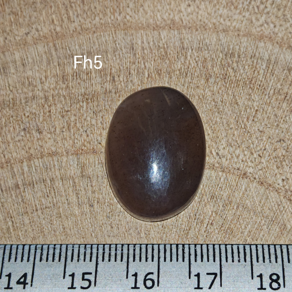Fekete holdkő-napkő kaboson, Fh5, ovális formájú, sötét alapszínű, milliméteres vonalzó mellett.”